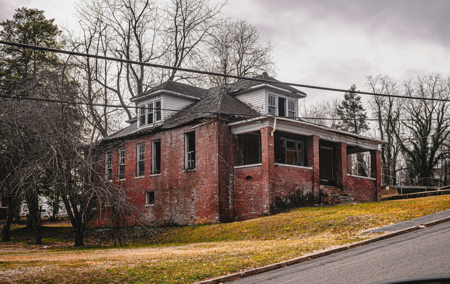 An old brick house with boarded-up windows and a weathered exterior, set against a cloudy sky, representing a home in need of repairs or an "as-is" property.